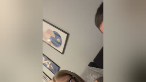 Media: A blurry video shows a gray wall with two framed prints of abstract, colorful figures, and a partially visible person in a gray sweater, possibly a child, standing in front.