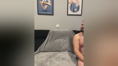 Media: A video of a man, partially visible, sitting on a gray-covered bed in a minimalist room with gray walls and two abstract artworks.