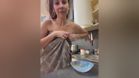 Media: Video of a light-skinned woman with shoulder-length hair, wrapped in a towel, standing in a kitchen. The background features shelves with various items, a sink, and a blue bowl.