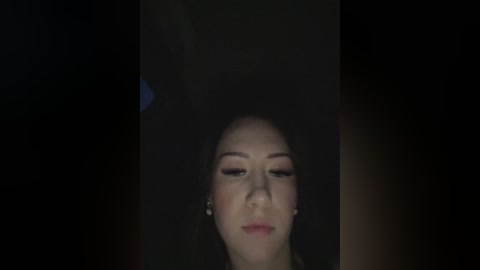Media: A dimly-lit video of a woman with fair skin, long dark hair, and subtle makeup, including eyeliner and pink lipstick, wearing small hoop earrings.