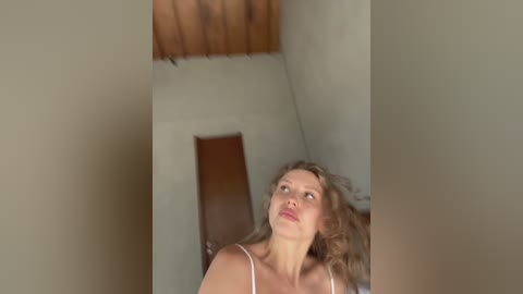 Media: Video of a Caucasian woman with light skin, curly blonde hair, wearing a white tank top, looking up with a pensive expression. She stands in a dimly lit, narrow room with beige walls and a wooden ceiling.