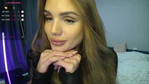 Video of a light-skinned woman with long brown hair, wearing a black top, sitting with hands clasped. Background includes a bed with white sheets and a dark curtain.