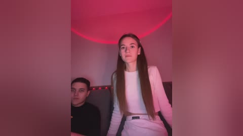 Media: Video of a woman in a white blazer and high-waisted pants, seated beside a man in a black shirt, both under a red-lit ceiling, creating a moody, minimalist ambiance.