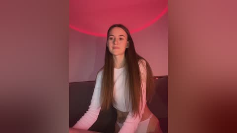 Media: Video of a young woman with long, straight brown hair, wearing a white long-sleeved top, sitting on a dark sofa. The background features a red spotlight.