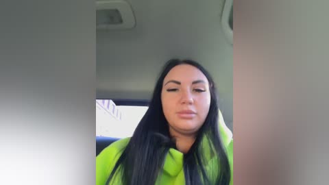 Video of a woman with long black hair, wearing a bright green jacket, sitting in a car. The background shows a blurry interior with a steering wheel and dashboard.