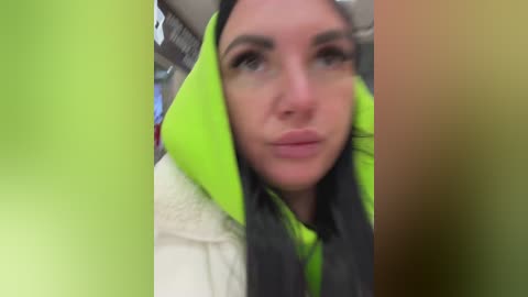 Video of a woman with long black hair, wearing a neon green hooded jacket and a white fur-trimmed coat, captured from a slightly blurry angle.