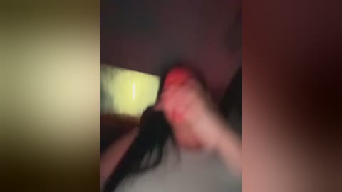 A blurred video showing a person with long black hair, red face, and hand to forehead, in a dimly lit room with yellow and red tones.