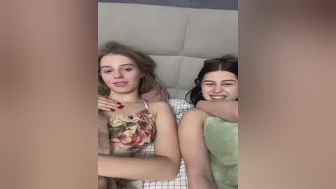 Media: Video of two young women, one with blonde hair in a floral top, the other with dark hair in a green shirt, cuddling in bed.