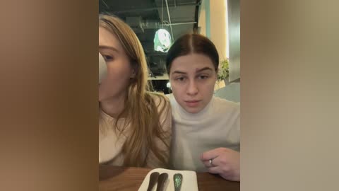 Media: Video of two women at a restaurant: a blonde woman with long hair and a brunette woman with a white turtleneck, both wearing neutral expressions.