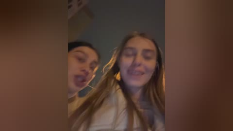 Media: Video of two young women with long hair, one in a black headband, the other in a white shirt, smiling and sticking out their tongues. Background is blurred.