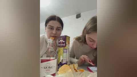 Media: Video of two young women, one with dark hair, the other with blonde, both wearing casual attire, eating snacks and looking at a bottle of Absolut vodka.