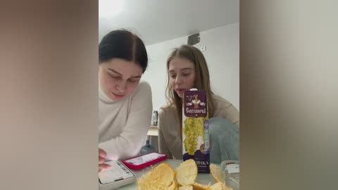 Media: A video of two young women with long hair, one in a white sweater, eating chips and drinking from a purple bottle at a cluttered kitchen table.