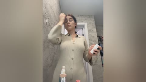Media: Video of a light-skinned woman with dark hair, wearing a beige button-up dress, applying makeup in a bathroom with a textured stone wall. She holds a white tube of concealer and a bottle of foundation.