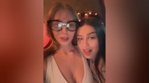 Media: A video of two women, one with glasses, both with fair skin, wearing low-cut white tops, smiling, taken from the side.