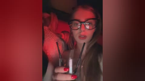 Media: Video of a woman with glasses, long blonde hair, and red nails, holding a lighter and cigarette, in a dimly lit room with red lighting.
