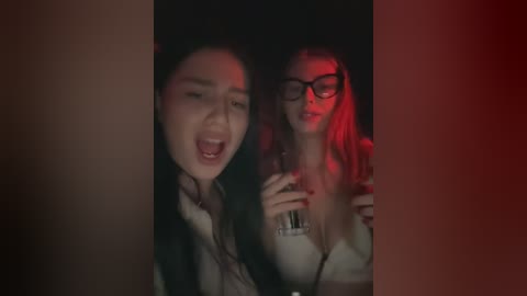 Media: Video: Two women, one Asian, one Caucasian, in dim lighting, laughing, holding glasses, red filter effect, intimate moment, suggestive.
