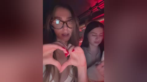 Media: Video of a young woman with long brown hair and glasses, making a heart shape with her fingers, standing next to a girl with long black hair, both in a dimly lit room with red lighting.