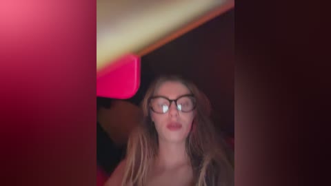 Media: A blurry video of a young woman with long blonde hair, wearing glasses, and a red dress, standing in a dimly lit room.