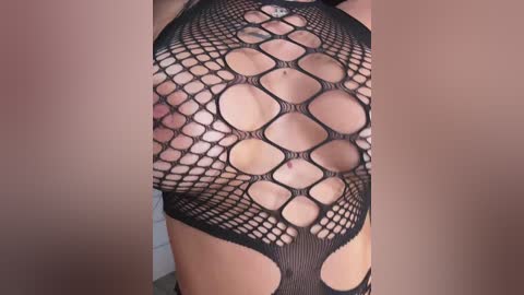 Media: Video of a woman wearing a black, fishnet bodysuit with diamond-shaped cutouts revealing her breasts and abdomen. The background is a plain beige wall.