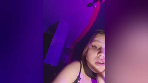 Video of a young woman with light skin, brown hair, and a black bra, leaning against a purple wall. She has a neutral expression, and the lighting casts a purple hue.