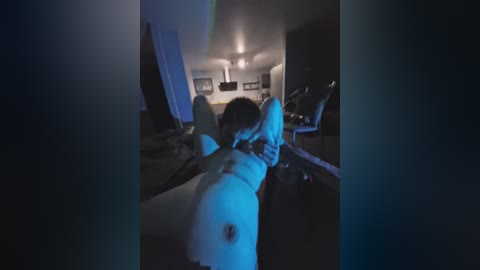 Video of a man with a shaved head, lying on a bed with his arms above his head, wearing a blue mask, in a dimly lit room with a ceiling fan.