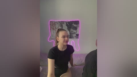 Video of a young, fair-skinned woman with a ponytail, wearing a black crop top and shorts, sitting on a bed with a gray wall behind her, featuring a neon heart outline and a poster of a dog's head.