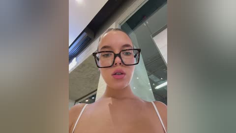 Media: Video of a young woman with fair skin, wearing black-rimmed glasses, a white spaghetti-strap top, and pink lipstick, taken from a low angle. Background shows modern office interior with glass walls and a skylight.