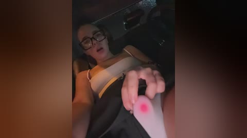 Media: Video of a woman in glasses, wearing a yellow and black striped top, holding a red object resembling a bloodied hand, in a dimly lit car.