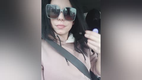 Media: A video of a young woman with long black hair, wearing large rectangular sunglasses and a light pink hoodie, driving a car.