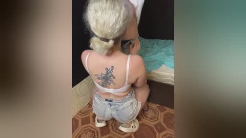 A video of a blonde woman with a braided ponytail, wearing a white bra and denim shorts, kneeling on a patterned rug. Her back displays a large, detailed tattoo of a butterfly.