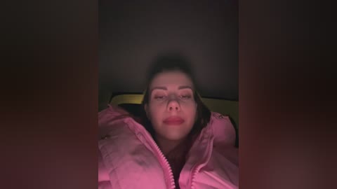 Media: Video of a young woman with pale skin, light brown hair, and closed eyes, lying in a dimly lit room, wearing a pink jacket.