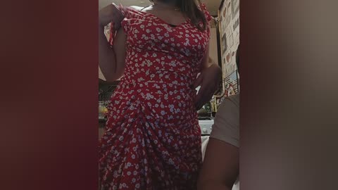 Video of a light-skinned woman in a red floral dress, holding it up, revealing a white thong, in a dimly lit, cluttered room with a brick wall and kitchen items visible in the background.