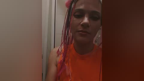 A video of a young woman with light brown skin and red hair extensions in a bathroom, wearing a bright orange tank top.