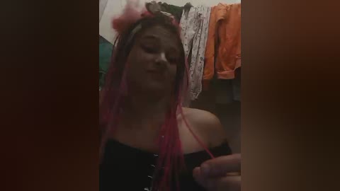 A video of a young woman with pink hair, wearing a black corset and headband, standing in a cluttered, dimly-lit room with orange and patterned clothes hanging behind her.