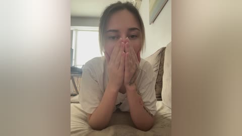 A young woman with light skin and straight, shoulder-length brown hair, wearing a white shirt, kneels on a beige carpet, hands clasped in prayer, in a dimly lit living room with wicker furniture and framed art on the wall.