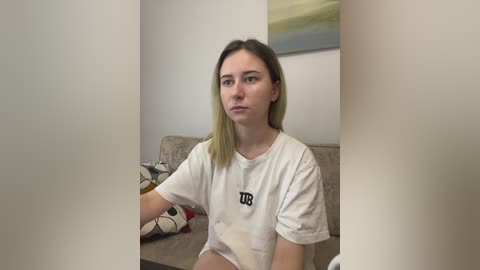 Video of a young Caucasian woman with straight, shoulder-length blonde hair, wearing a white T-shirt with the word \"UB\" on it, sitting on a beige couch in a minimalist room with a painting on the wall.