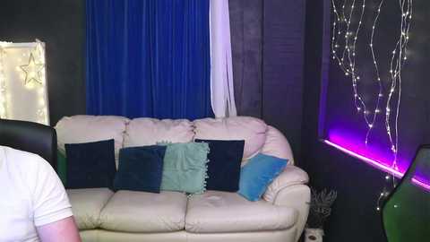 Video of a dimly lit living room with a white leather couch adorned with dark blue and teal pillows, dark blue curtains, a white wall, and glowing fairy lights.