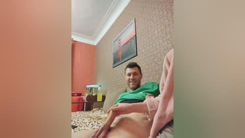 Media: A video of a middle-aged man with a beard, wearing a green shirt, seated on a bed with leopard print bedding, in a room with patterned wallpaper and a framed painting.