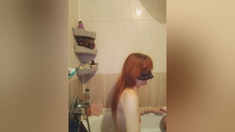 A video of a nude, fair-skinned redhead woman with long hair, wearing a black blindfold, sitting in a bathtub with beige tiles and a shower caddy filled with toiletries.