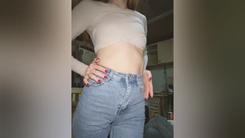 Video of a light-skinned woman with long brown hair, wearing a cropped, ribbed, white crop top and high-waisted, blue jeans. She has red-painted fingernails. The background shows a dimly lit, cluttered room with wooden furniture and a TV.