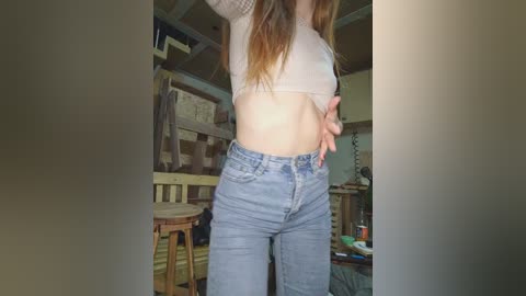 Video of a young woman with long brown hair, wearing a white crop top and high-waisted blue jeans, standing in a cluttered bedroom with wooden furniture and a messy bed in the background.