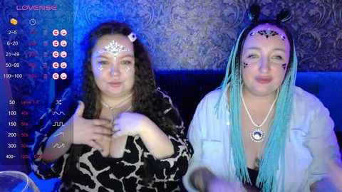 Video of two women with elaborate makeup, wearing vibrant clothing, sitting at a table. One has curly hair, the other turquoise braids, with a blue backdrop and a \"LOVENICE\" overlay.
