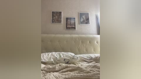 Media: Video of a minimalist bedroom with a cream-colored tufted headboard, three framed pictures on a beige wall, and a disheveled white bed with a plush blanket.