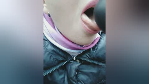 A close-up video of a person's mouth, tongue, and upper chest, wearing a black puffer jacket over a pink and white striped shirt. The image is blurred with a gradient background.