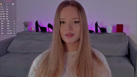 Video of a young Caucasian woman with long, straight blonde hair, wearing a white sweater, sitting on a gray couch in a modern, softly lit living room with purple ambient lighting and black high-heeled shoes displayed on a shelf behind her.