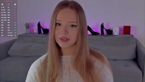 Video of a young, fair-skinned woman with long, straight blonde hair, wearing a white sweater, sitting on a gray sofa in a modern living room with purple lighting and black high heels on the backrest.