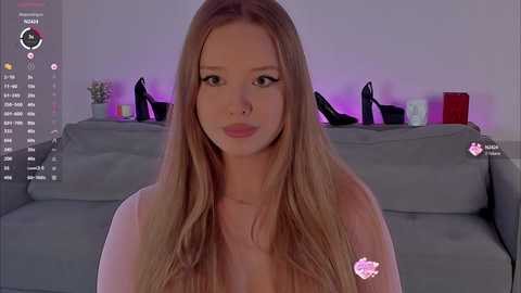 Video of a blonde, fair-skinned woman with long hair and light makeup, sitting on a gray couch. Black heels and candles on a shelf in the background.