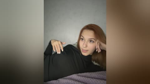 Media: A video of a young Asian woman with straight, light brown hair, wearing a black turtleneck, lying on a bed with a purple blanket, resting her head on her hand against a textured, grey wall.