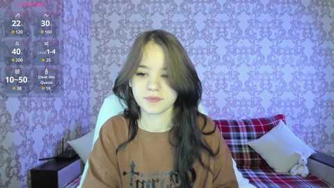 Media: A video of a young girl with long, wavy brown hair, sitting on a bed with red and white checkered sheets, in a room with floral wallpaper. She wears a brown shirt.