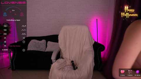 Video of a woman, partially covered by a white blanket, sitting on a black couch, with pink lighting and a Halloween-themed screen in the background.